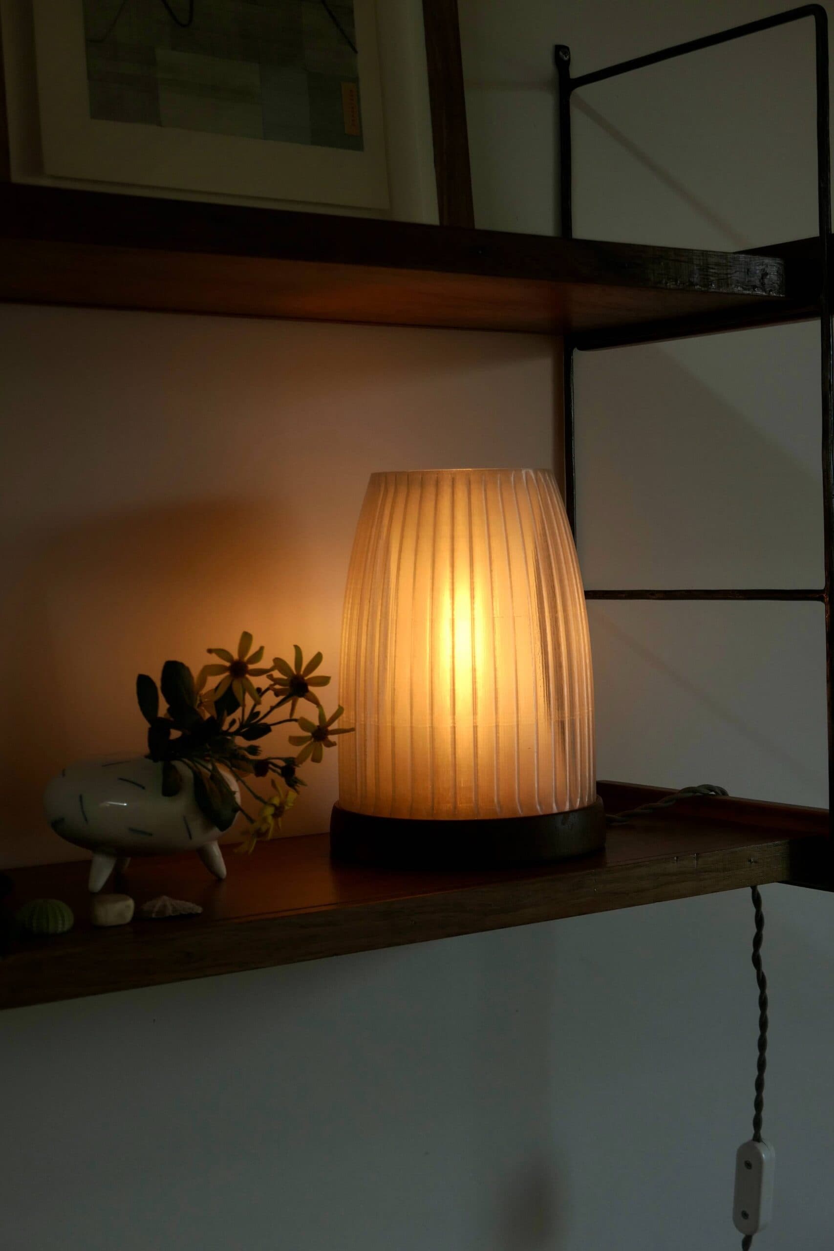 Fault Lines Table Lamp switched on, warm light through the fluted shade on a wooden shelf